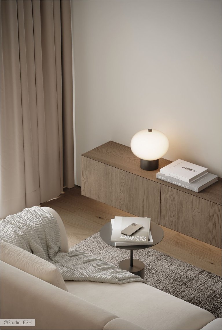 Интерьер гостиной с белым диваном, серым ковром и деревянной мебелью. На журнальном столике лежат книги, а на тумбе стоит лампа. В комнате много естественного света