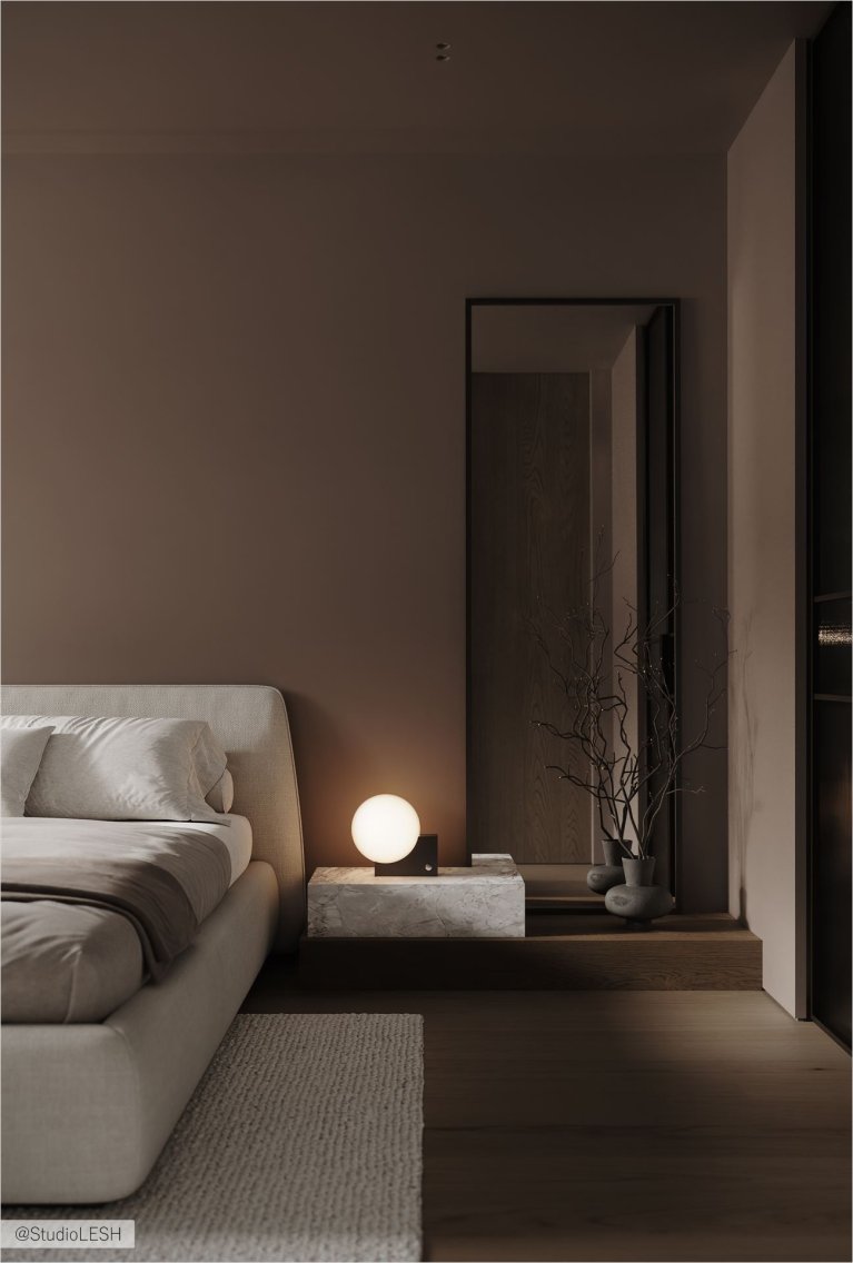 Уютная спальня с большой кроватью, уставленной мягкими подушками и покрытой покрывалом. На прикроватной тумбочке стоит лампа, рядом висит зеркало в полный рост. В комнате преобладают теплые оттенки коричневого и бежевого