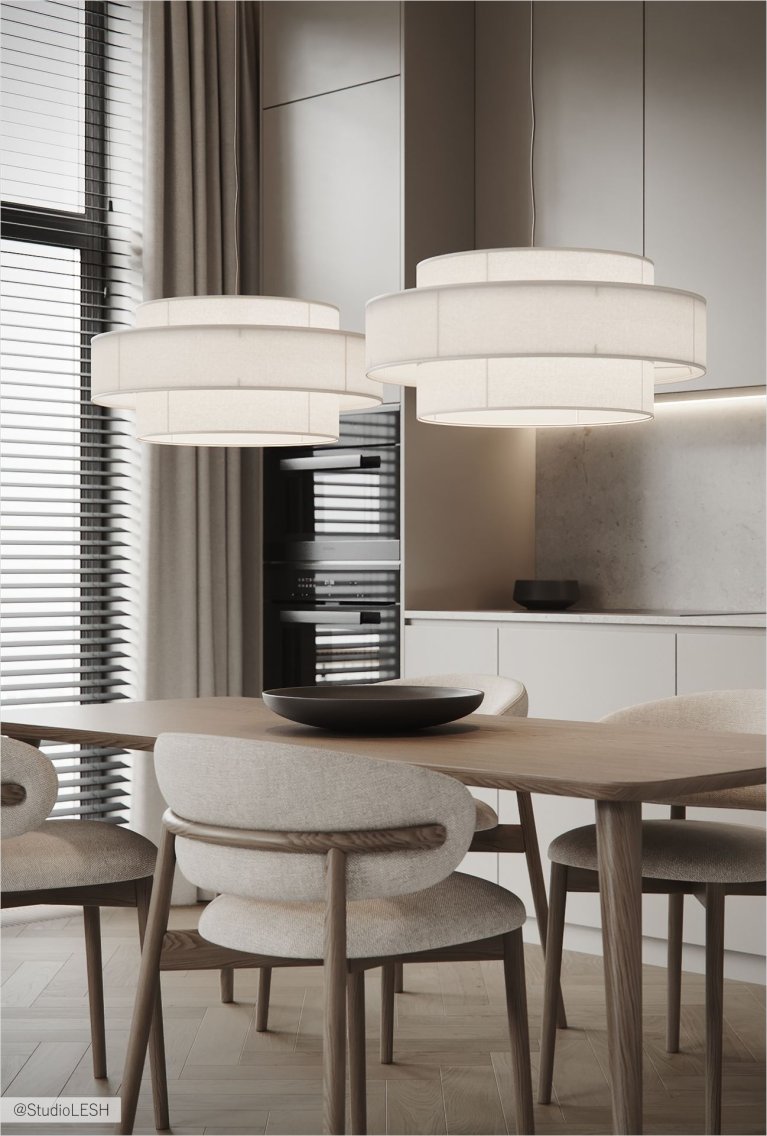 Интерьер кухни в светлых тонах с минималистичными фасадами, встроенной техникой, деревянным обеденным столом и четырьмя стульями. Над столом висят два белых подвесных светильника