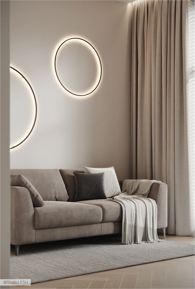 Просторная гостиная с серым диваном, украшенным подушками, и двумя круглыми настенными светильниками. В комнате также есть высокие окна с плотными шторами, паркетный пол и серый ковер