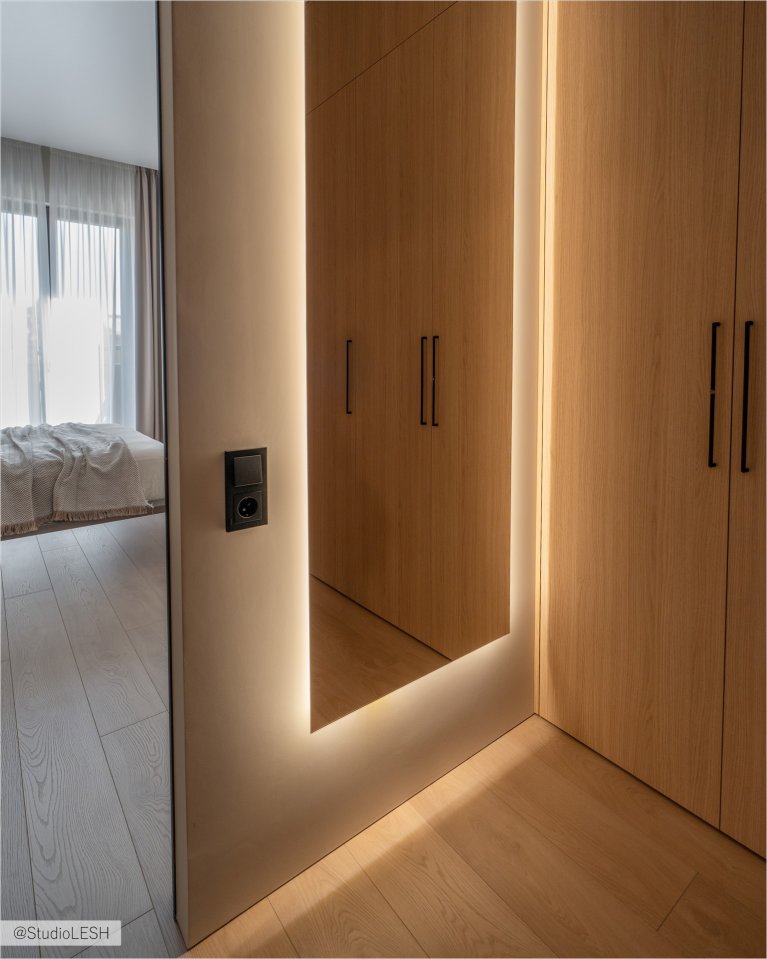 Стильный коридор с большим зеркалом, визуально увеличивающим пространство. Справа - встроенный шкаф с гладкими деревянными фасадами и минималистичными ручками. На полу - светлый паркет, создающий уютную атмосферу
