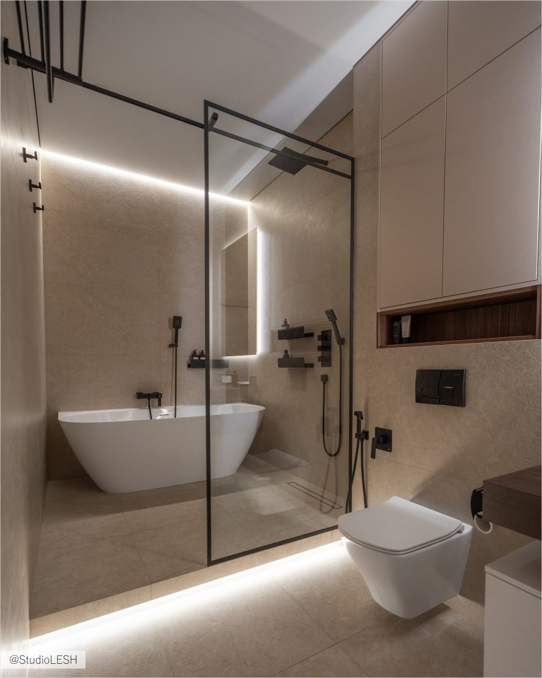 Стильная ванная комната с душевой кабиной с тропическим душем и стеклянной перегородкой. Слева - отдельно стоящая ванна с черным смесителем. Справа - подвесной унитаз и встроенные шкафы с подсветкой. Вся комната выполнена в светлых тонах с использованием натуральных материалов