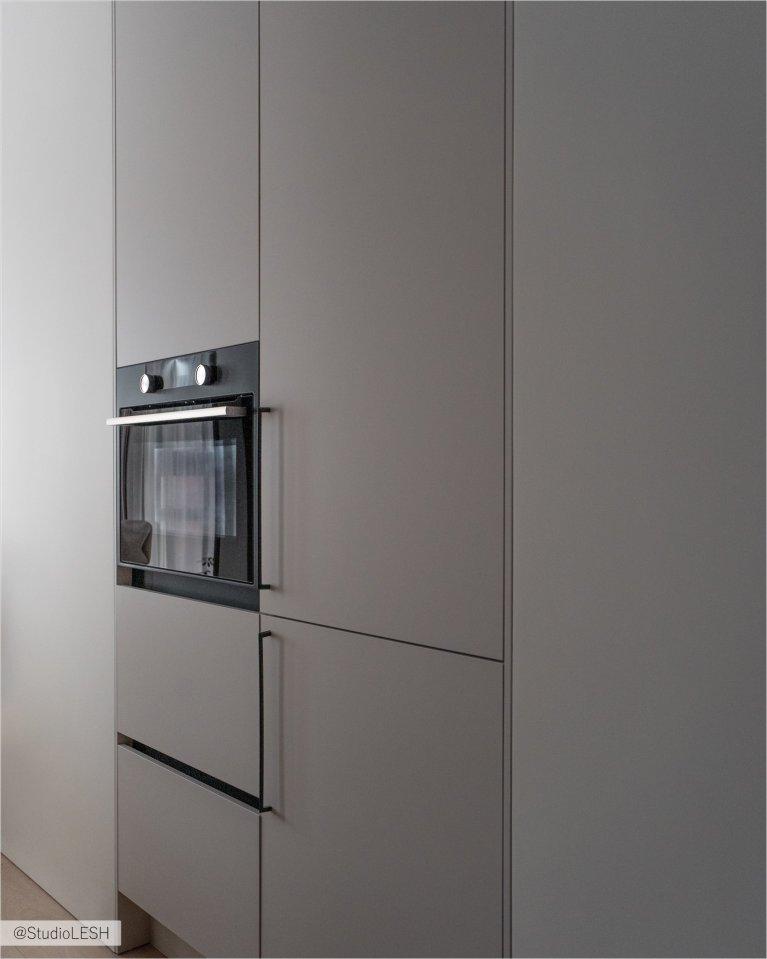 Интерьер кухни в светлых тонах с гладкими фасадами, встроенным духовым шкафом с сенсорной панелью управления, и минималистичными ручками. Видны также другие элементы кухонного гарнитура, выполненные в едином стиле