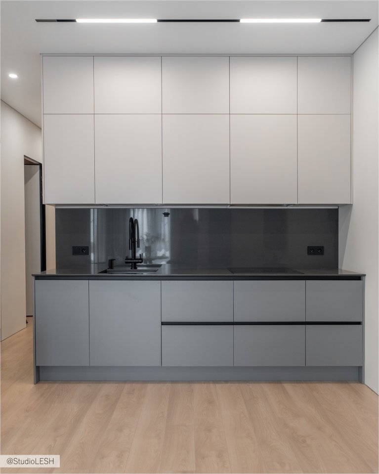 Интерьер кухни в светлых тонах с гладкими фасадами, встроенным духовым шкафом с сенсорной панелью управления, и минималистичными ручками. Видны также другие элементы кухонного гарнитура, выполненные в едином стиле