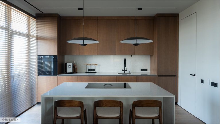 Стильная кухня с деревянными шкафами, белым кухонным островом с встроенной плитой и тремя современными стульями. Над островом висят два минималистичных светильника. Окно с жалюзи обеспечивает естественное освещение
