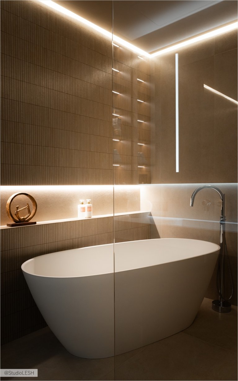 Стильная ванная комната с отдельно стоящей ванной, оборудованной современным смесителем. На заднем плане - душевая кабина со стеклянной перегородкой и полочки с банными принадлежностями. Освещение играет важную роль в создании атмосферы: подсветка под ванной и на стенах подчеркивает текстуру плитки и создает уют