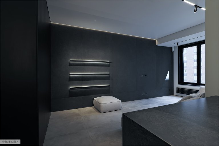 Стильная гостиная с серым диваном, украшенным подушками, и темным столиком. На заднем плане - большое окно с видом на город, черная стена с подсветкой и минималистичные светильники, создающие уютную атмосферу