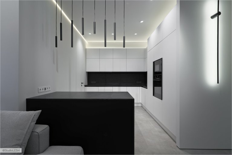 Стильная кухня с белыми шкафами, черным кухонным островом с темной столешницей, над которым висят минималистичные светильники. Встроенная техника гармонично сочетается с общим дизайном кухни. Освещение играет важную роль в создании уютной атмосферы