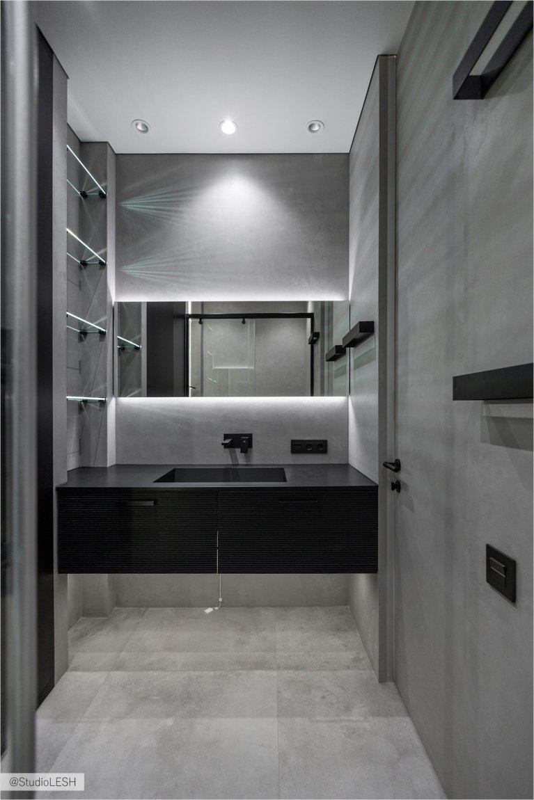 Стильная ванная комната с подвесной раковиной, установленной на столешнице из темного дерева. Над раковиной висит большое зеркало с подсветкой, отражающее светлые стены и полки с аксессуарами. Видны также черные смесители и электрические розетки