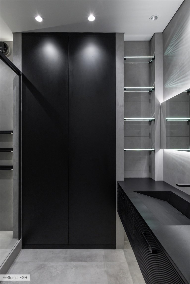 Стильная ванная комната с темными шкафами с гладкими фасадами, подчеркнутыми встроенной подсветкой. Подсветка создает игру света и тени, делая интерьер более выразительным. Минималистичный дизайн ванной комнаты подчеркивает ее современность и функциональность