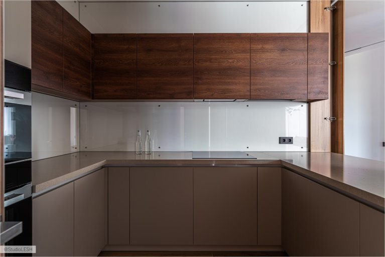 Современная U-образная кухня с деревянными фасадами, белой столешницей и стильным освещением